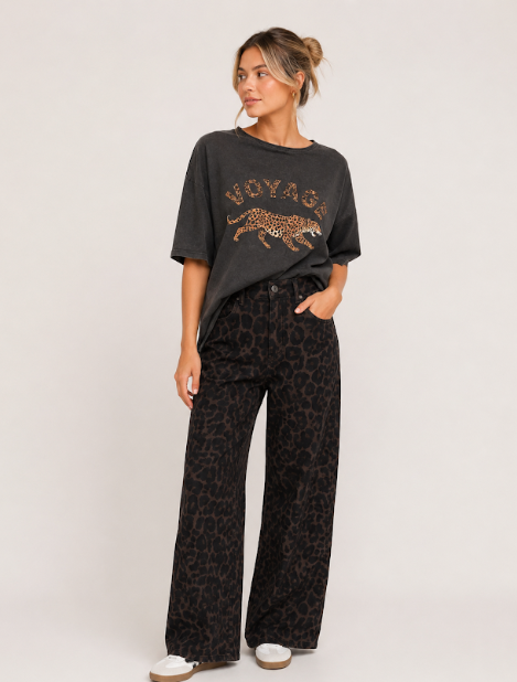 A woman modeling a dark charcoal gray t-shirt with a leopard print graphic and the word 'NOYAGE' on the front, paired with black leopard print wide-leg pants and white slip-on shoes, against a plain light gray studio background.