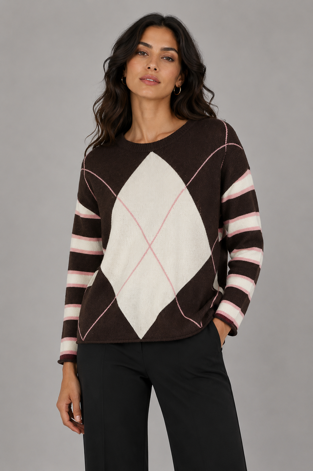 A woman modeling a long-sleeve knit sweater with a brown and cream argyle pattern and thin pink intersecting lines, paired with black pants against a solid gray studio background.