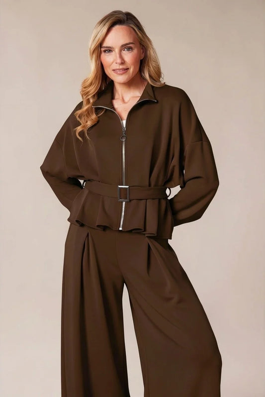 A woman modeling a dark brown two-piece jumpsuit with a zip-up long-sleeve top and matching wide-leg pants, set against a plain beige studio background.