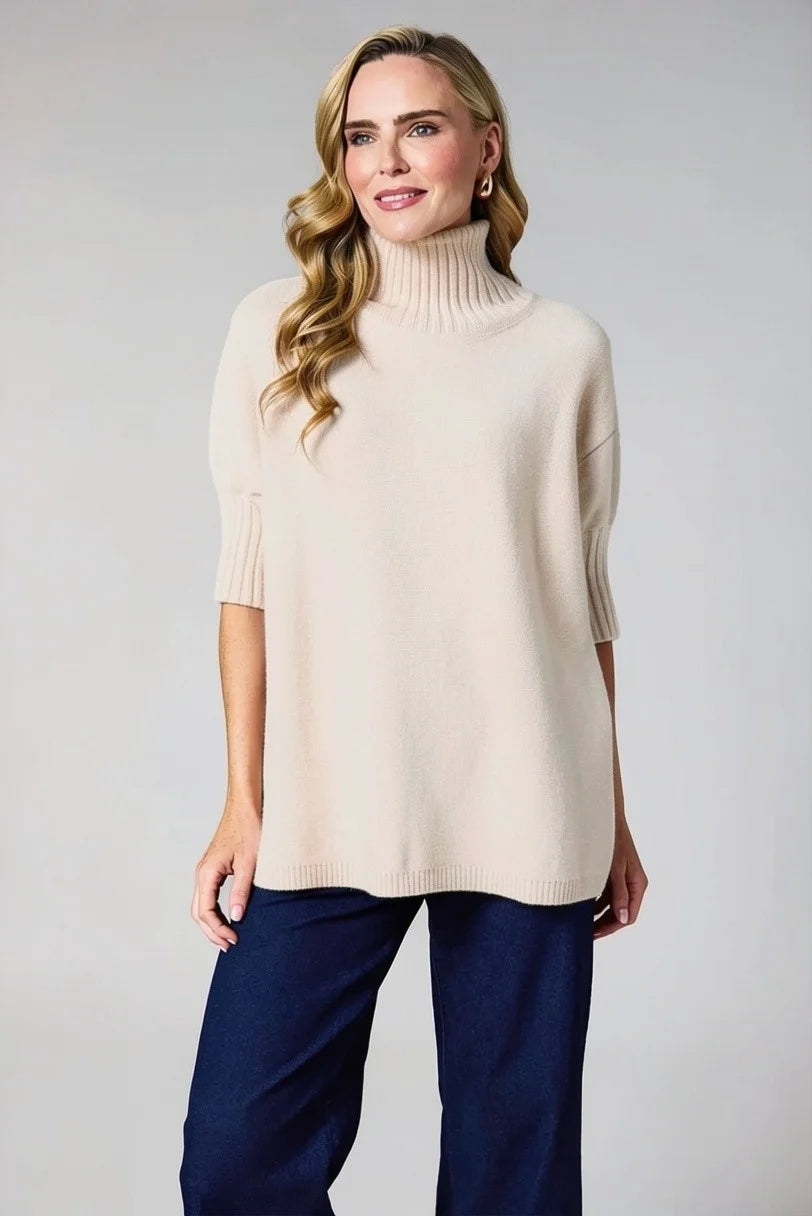 A woman modeling a cream-colored knit turtleneck sweater with elbow-length sleeves, paired with dark blue pants, against a solid light gray background.