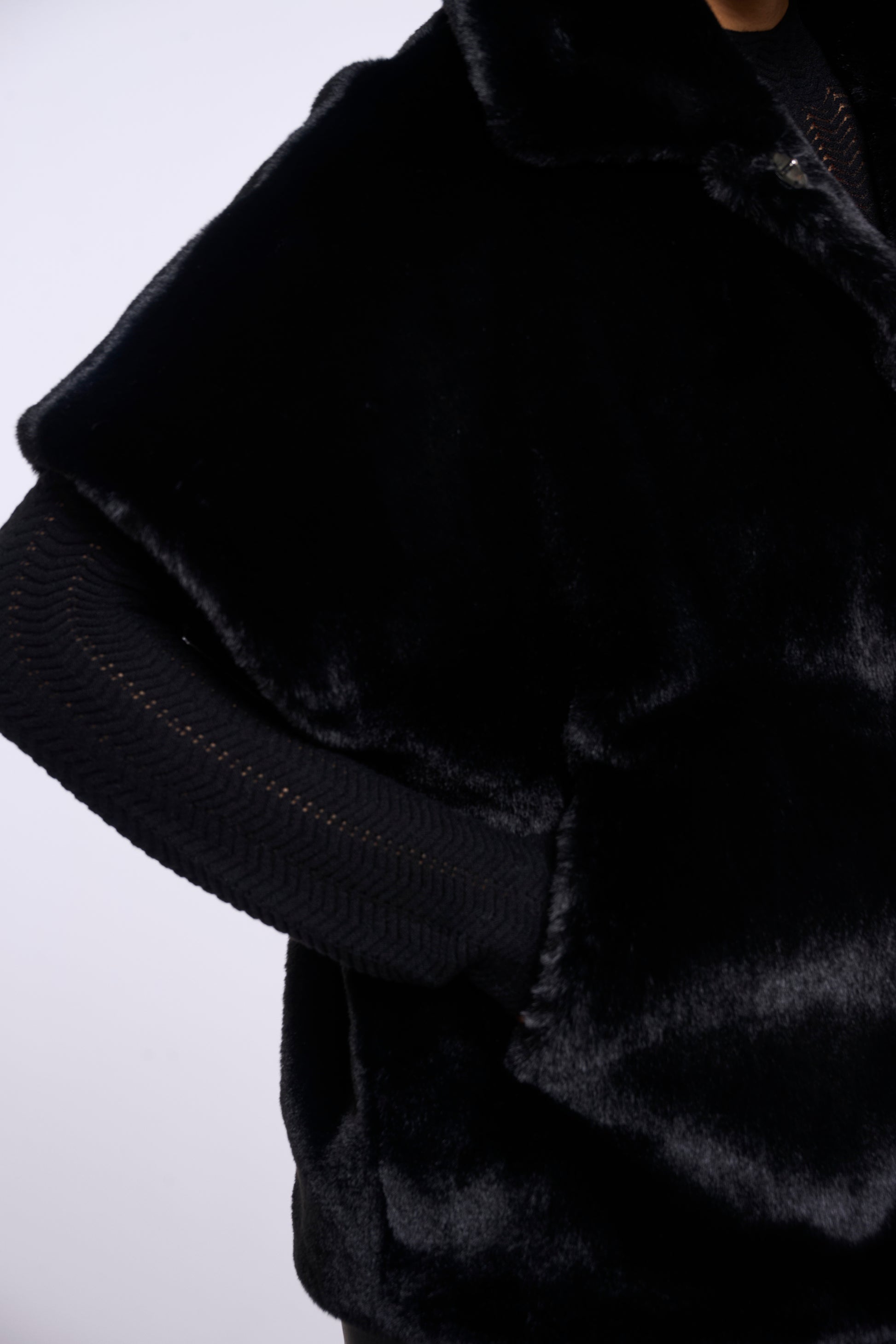 Close-up of a black faux fur jacket with a ribbed knit sleeve and collar, shown on a mannequin against a light gray background.
