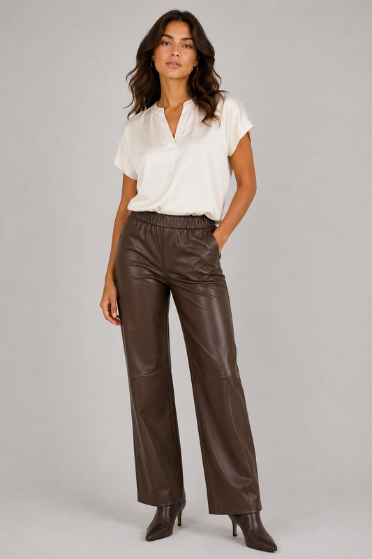 A woman modeling a white short-sleeve blouse and dark brown wide-leg faux-leather pants against a plain light gray studio background.
