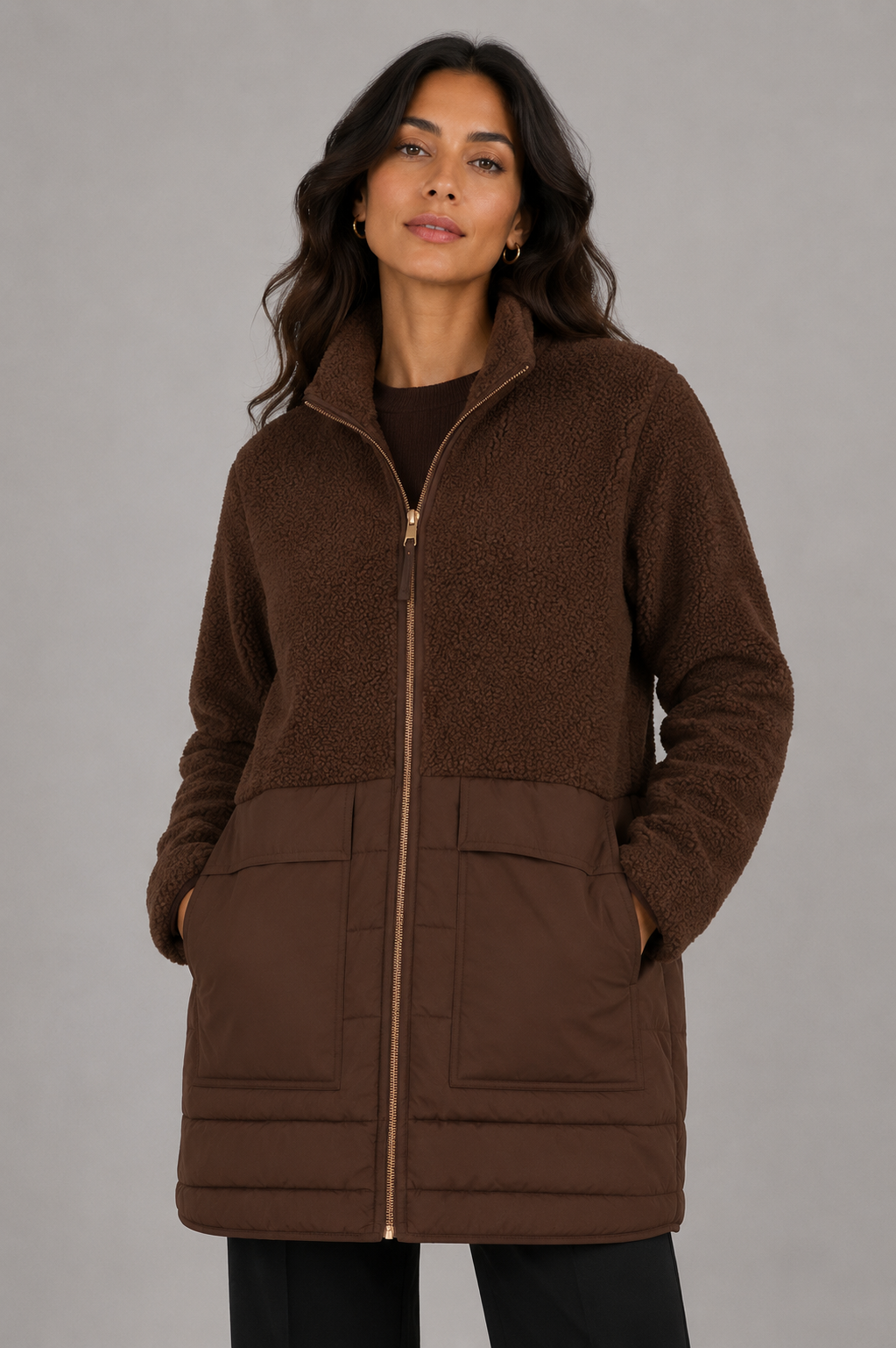 A woman wearing a brown fleece zip-up jacket with a quilted lower panel and front pockets, photographed against a plain gray studio background.
