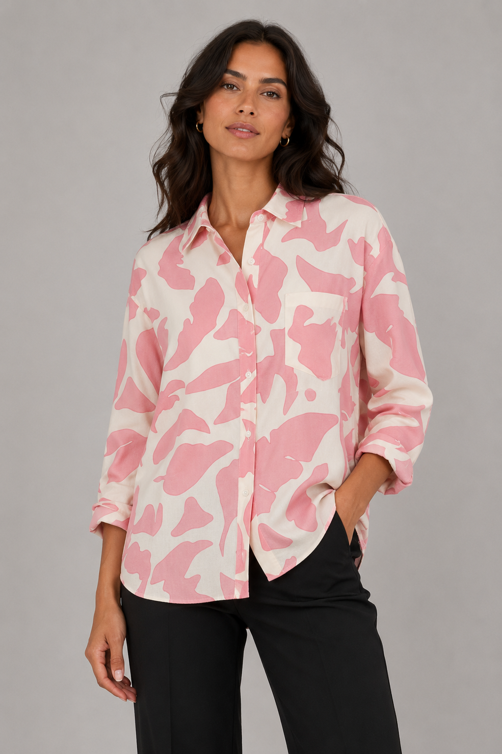 A woman modeling a pink and off-white abstract patterned button-up shirt with black pants, photographed against a solid gray studio background.
