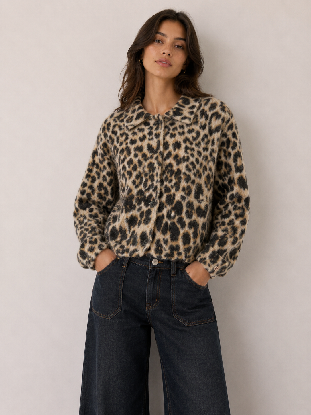 A woman wearing a beige and black leopard print fuzzy zip-up jacket with dark wide-leg jeans, standing against a plain light gray wall.